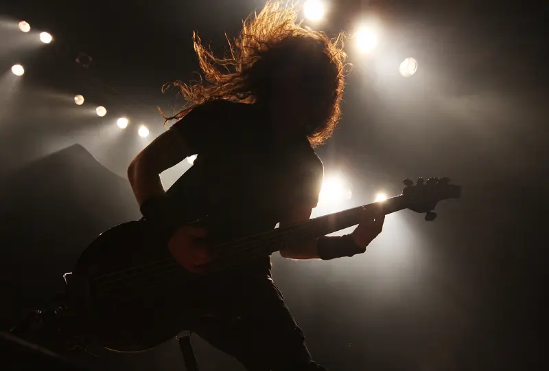 Silhouetted musician with flowing hair playing bass guitar on stage, surrounded by bright stage lights.
